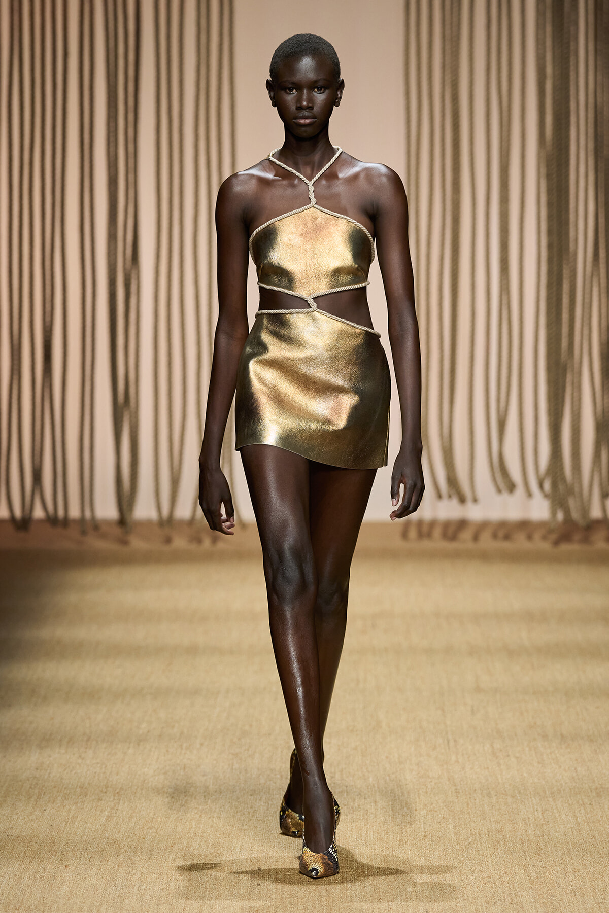 Model walks the runway in a gold metallic two-piece outfit with rope halter and wrap details on a fashion show runway.