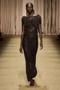 Model walking runway in a dark, intricately woven brown crochet gown with fringe shoulders.