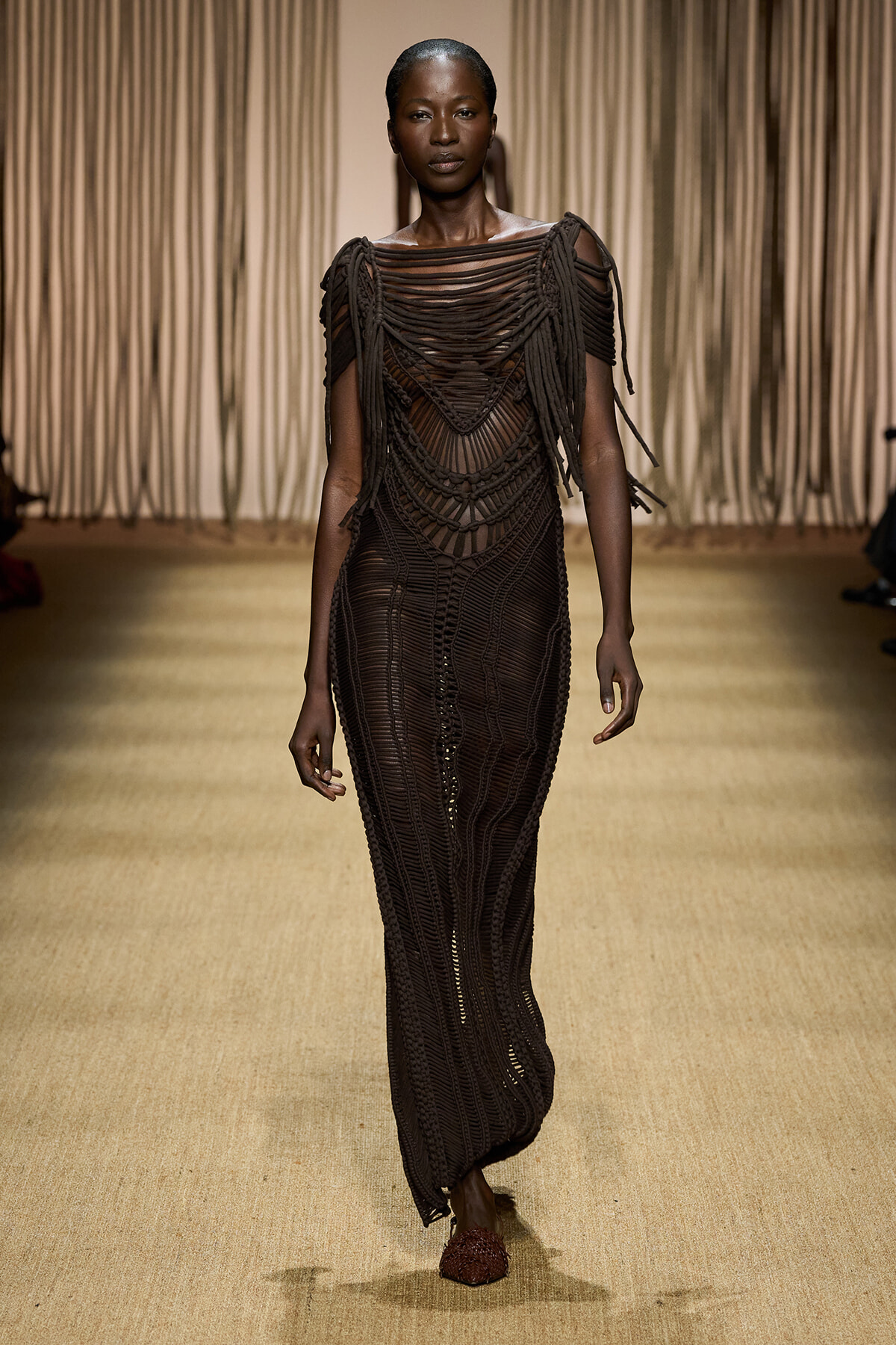 Model walking runway in a dark, intricately woven brown crochet gown with fringe shoulders.