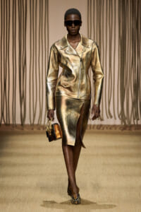Model walking the runway in a metallic gold leather jacket and matching gold skirt with a front slit, dark sunglasses, and snake-print heels, carrying a small crocodile-patterned handbag
