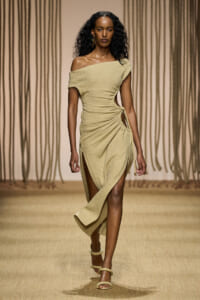 Model walking the runway in a beige off-shoulder textured dress with a side tie and high thigh slit, gold earrings, and braided sandals under warm lighting.