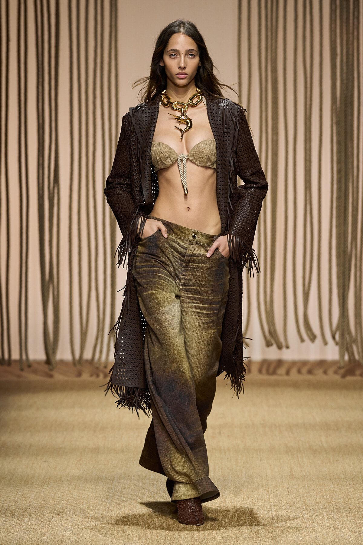 Model walks the runway in a beige bandeau top, dark fringed coat, and olive metallic pants, accessorized with a chunky gold chain necklace.