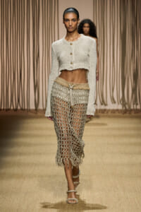 Model walking the runway in a cream cropped knit cardigan with gold buttons and a long, open-weave crochet skirt paired with strappy sandals.