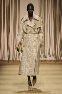 Model walks the runway in a long beige textured trench coat with gold buttons and a rope-strap bag.