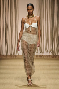 Model walks the runway in a cream bandeau top with a beige crochet mesh dress and a chunky gold necklace, paired with rope sandals.