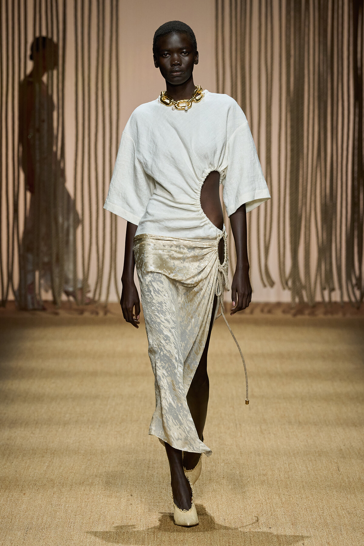 Model walks the runway in a white oversized top with a side cut-out and a draped, metallic-cream skirt with a knot detail and rope accent, gold chunky necklace.