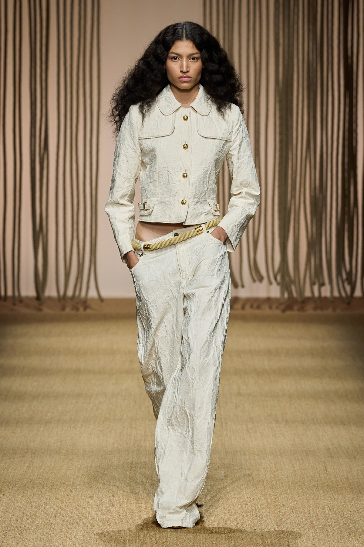 Model on a runway wearing a crinkled cream jacket and wide-leg pants with gold buttons and a rope belt.