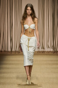 Model walks the runway in a white textured bikini top and matching mid-length skirt with rope accents, beige backdrop behind.