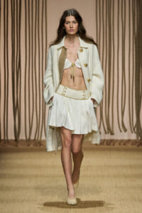 Model walks the runway in a cream coat, matching high-waisted shorts, and a white bikini top with a gold-tasseled front detail; beige backdrop.