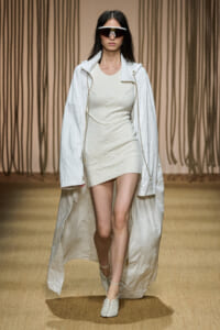 Model on runway wearing a white textured mini dress with a long crinkled white coat and futuristic white goggles on a beige backdrop.