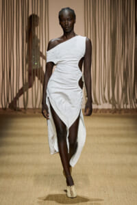 Black model walking a runway in a white one-shoulder, asymmetrical dress with side cutouts and a thigh-high hemline on a beige catwalk.