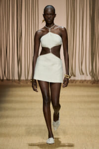 Black female model walking the runway in a white cutout halter dress with rope trim and side cutouts, paired with woven shoes and chunky gold bracelets.