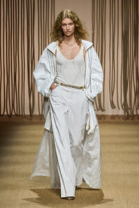 Model walks a fashion runway wearing an all-white ensemble: oversized jacket over a knit bodysuit and wide-leg pants with a rope belt and gold accents.