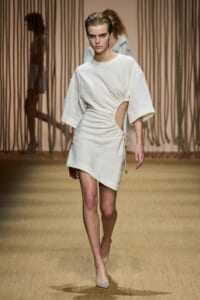 Model walking the runway in a white, knee-short dress with a side cutout and gathered waist detail; neutral-toned background.