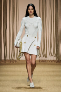 Model walking runway in a white knit top with geometric stitching and a high-waisted white mini skirt with gold buttons; carrying a white bag with a thick rope strap and wearing white heeled sandals.