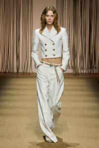 Model walks runway in an ivory cropped double-breasted blazer with gold buttons and matching high-waisted wide-leg pants, white rope belt.