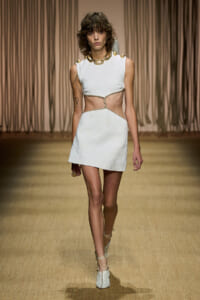 Model walks the runway in a white textured mini dress with waist cutouts, paired with a chunky gold necklace and white heeled sandals.