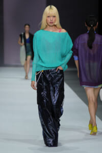 Model walks the runway in a teal knit off-shoulder sweater and glossy black cargo pants, with a blonde bob hairstyle.