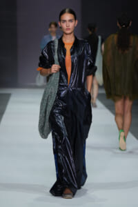 Runway model wearing a dark glossy jumpsuit with an orange top underneath, carrying a glittery silver tote on her shoulder.