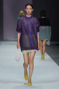 Model walks the runway in a sheer purple oversized top over lime shorts, yellow flats, and a white textured handbag.