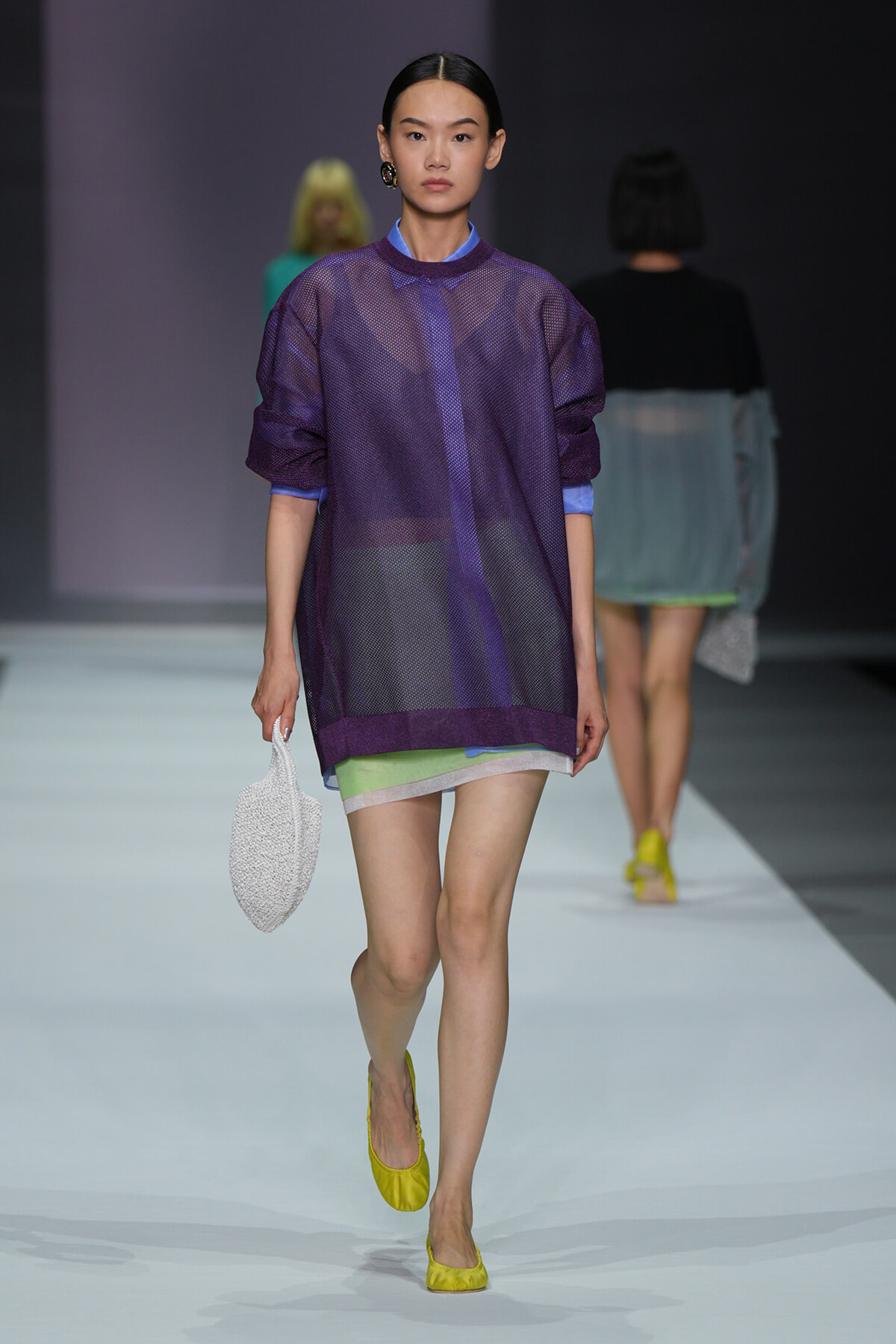 Model walks the runway in a sheer purple oversized top over lime shorts, yellow flats, and a white textured handbag.