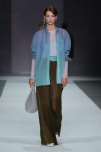 Female model walking a runway in a light blue overcoat over pastel knit top and wide brown pants, holding a sparkly silver bag.