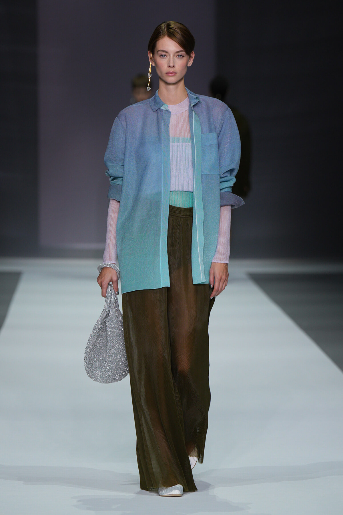 Female model walking a runway in a light blue overcoat over pastel knit top and wide brown pants, holding a sparkly silver bag.