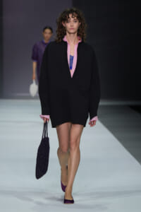 Model walks a fashion runway wearing an oversized black coat with pink cuffs and pink collar, layered over a purple top, carrying a black beaded tote and wearing purple flats.