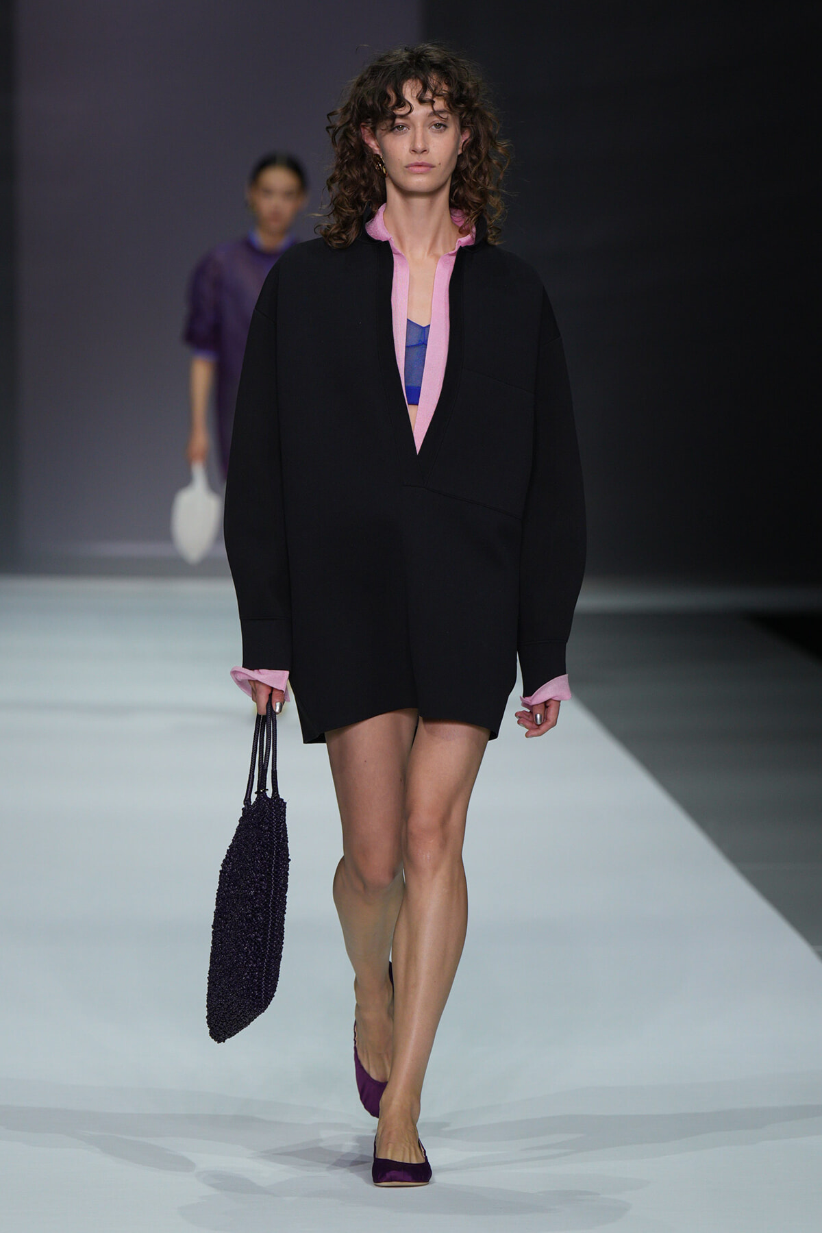 Model walks a fashion runway wearing an oversized black coat with pink cuffs and pink collar, layered over a purple top, carrying a black beaded tote and wearing purple flats.