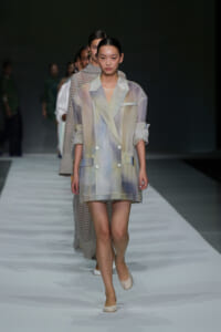 Model walking the runway in a pastel, double-breasted blazer dress with rolled sleeves and large pockets (soft, sheer fabric). Use of light, washed colors with white buttons.