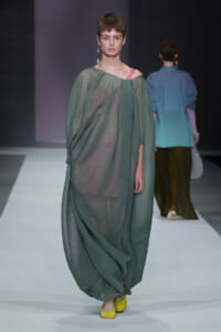 Model walks a runway in a loose, sheer emerald-green gown with a pink strap showing at the shoulder and yellow flats.