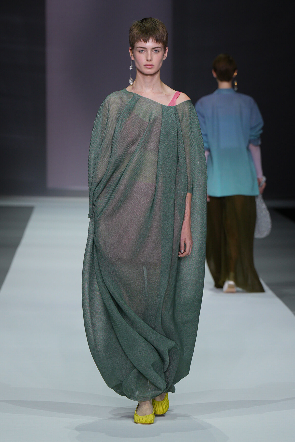 Model walks a runway in a loose, sheer emerald-green gown with a pink strap showing at the shoulder and yellow flats.