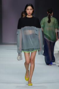 Female model walking a runway in a two-tone top (black yoke with sheer gray-blue body) over lime shorts, bright yellow flats, and a sparkling silver clutch; minimalist backdrop indicates a fashion show.