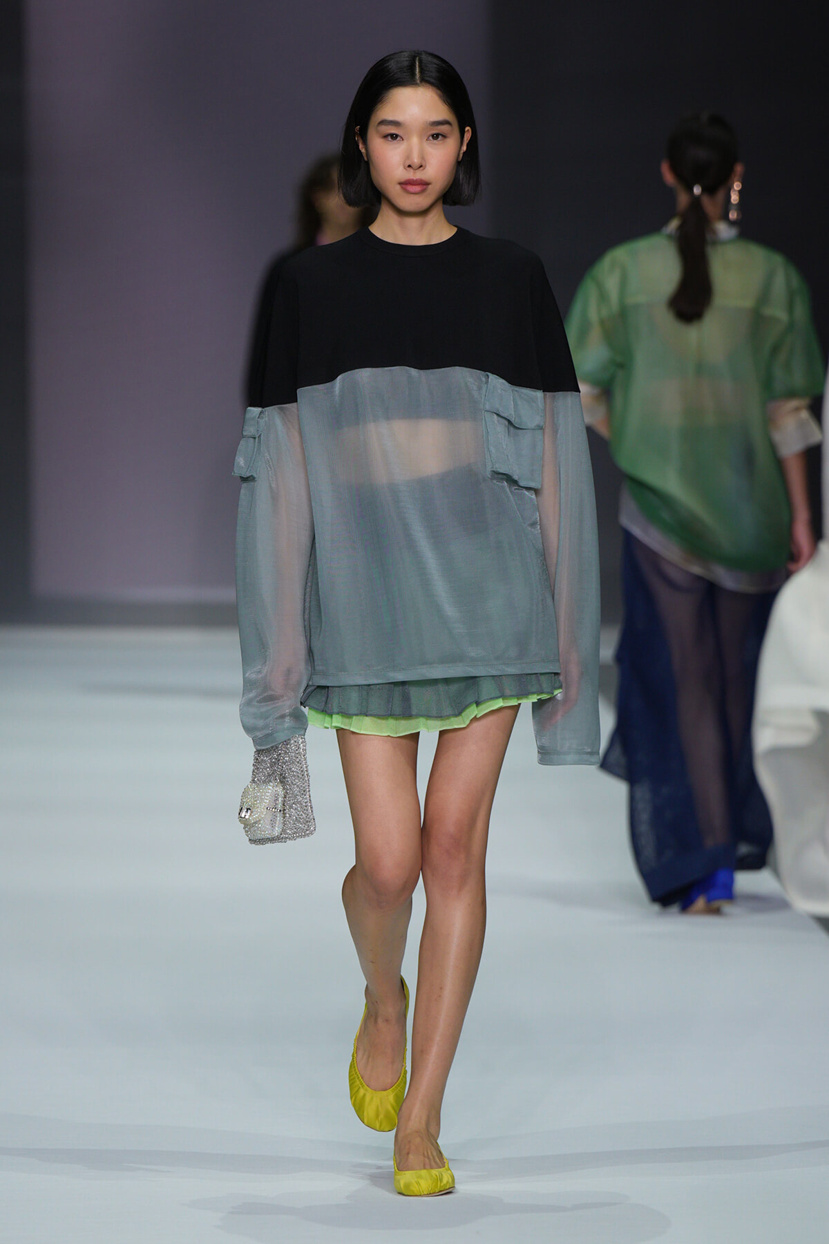 Female model walking a runway in a two-tone top (black yoke with sheer gray-blue body) over lime shorts, bright yellow flats, and a sparkling silver clutch; minimalist backdrop indicates a fashion show.