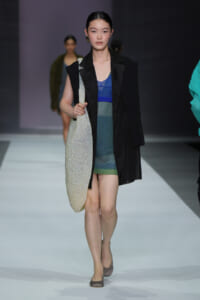 Model walking a fashion runway with a glittery large tote bag over her shoulder, wearing a layered blue-green dress and black outerwear.