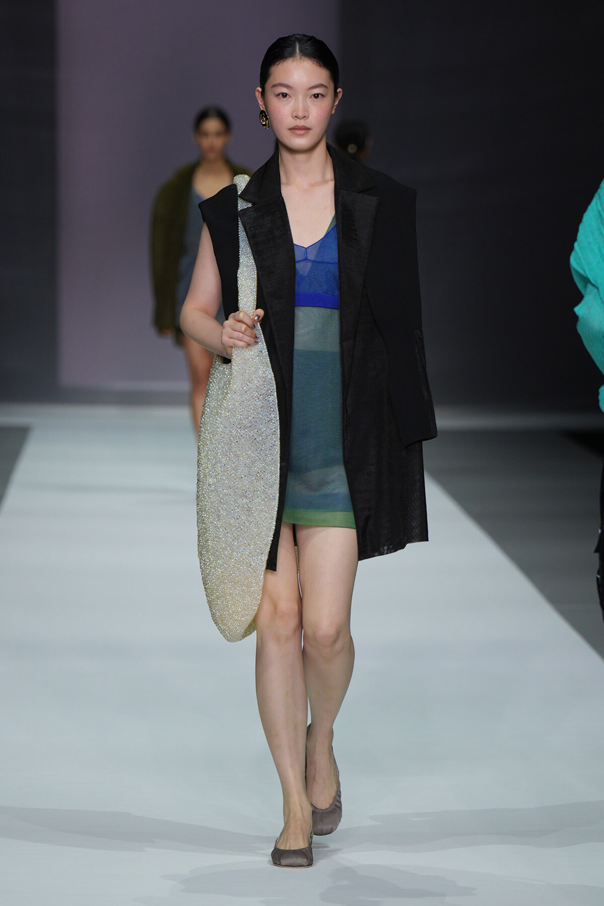 Model walking a fashion runway with a glittery large tote bag over her shoulder, wearing a layered blue-green dress and black outerwear.