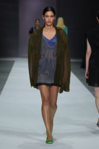Model walks the runway in a sheer blue-gray slip dress with embroidered floral motifs, layered under a loose olive-green sheer cardigan and bright green heels.