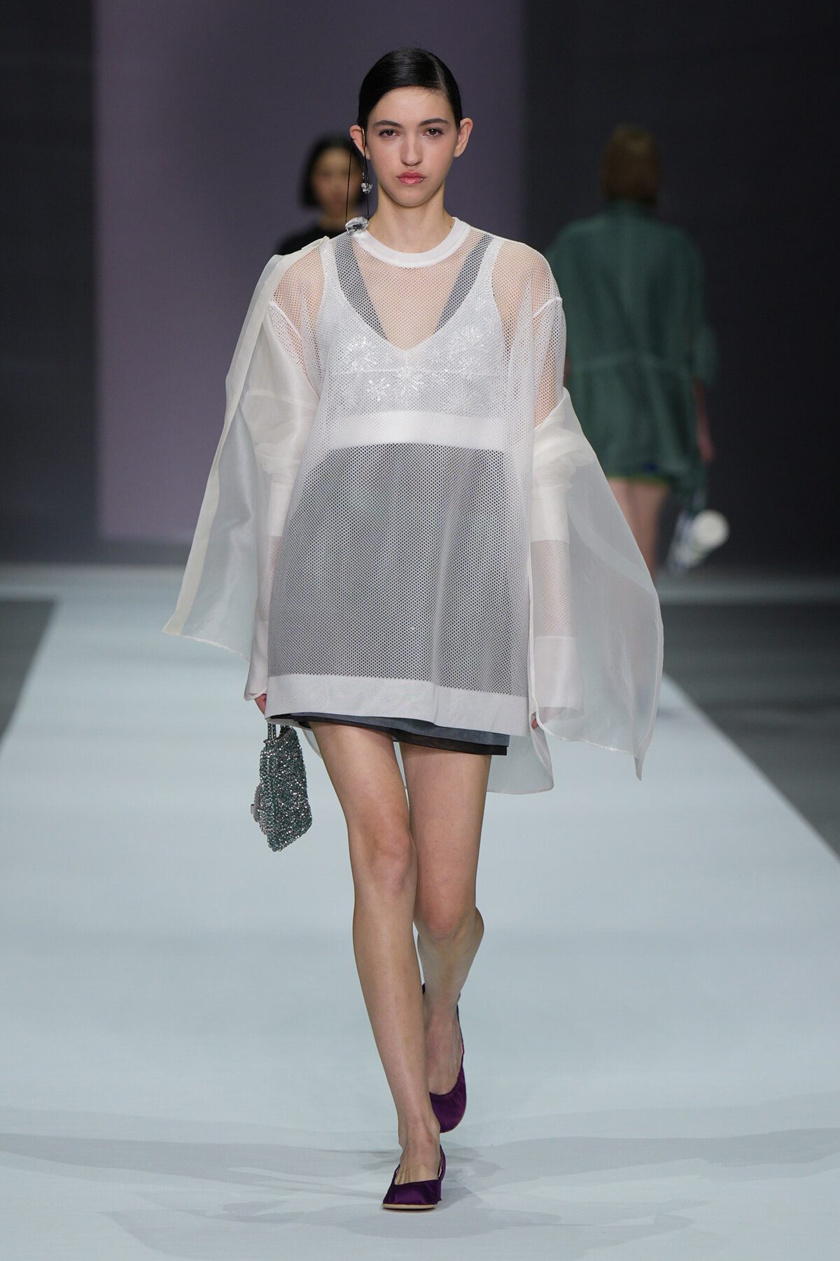 Model walks a fashion runway in a sheer white mesh dress with layered undergarments, holding a beaded bag and wearing purple flats