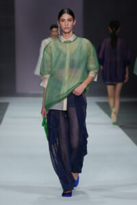 Model walking the runway in a sheer green top layered over a light shirt, with navy mesh pants and blue shoes, carrying a green bag.