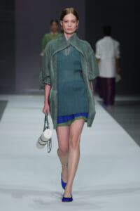 Model walking a runway in a teal knit mini dress with a loose green cape, carrying a small white circular handbag and wearing blue flats; soft studio backdrop.