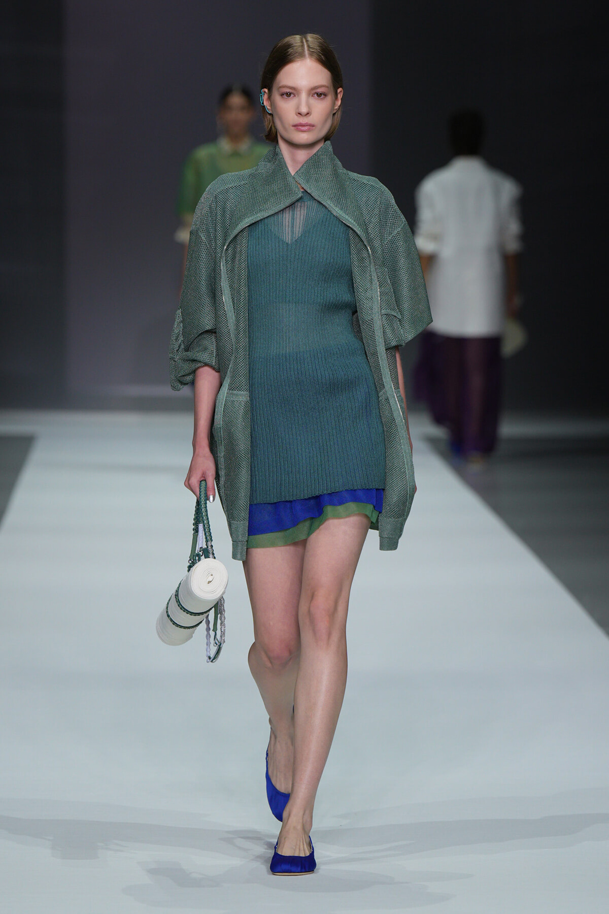 Model walking a runway in a teal knit mini dress with a loose green cape, carrying a small white circular handbag and wearing blue flats; soft studio backdrop.