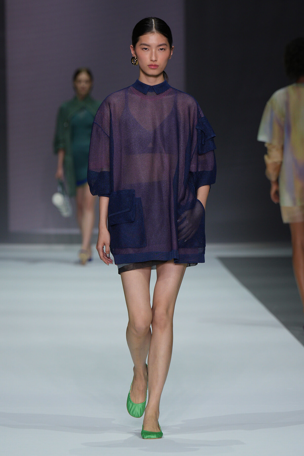 Model walking the runway in a sheer navy-purple tunic with oversized pockets and a dark collar, paired with green flats.