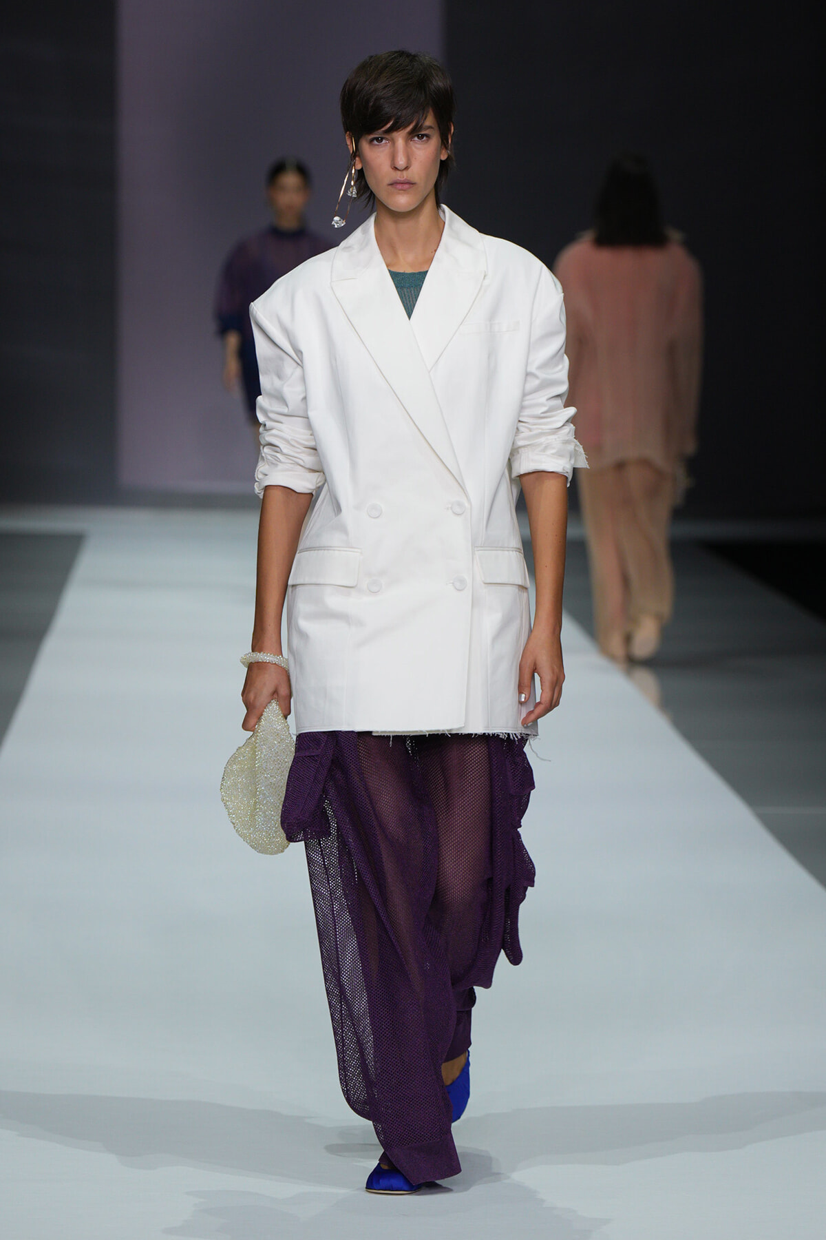 Model walks the runway in a white double-breasted blazer over a sheer purple skirt, holding a beaded bag at her side.