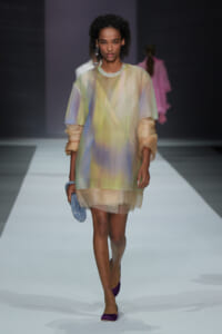 Model walking a runway in a pastel sheer shift dress with layered lining, holding a blue beaded clutch and wearing purple flats.