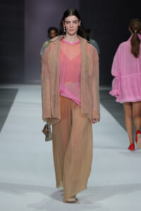 Model walks runway in peach sheer shirt and oversized beige outerwear with wide beige trousers, holding a beaded clutch.