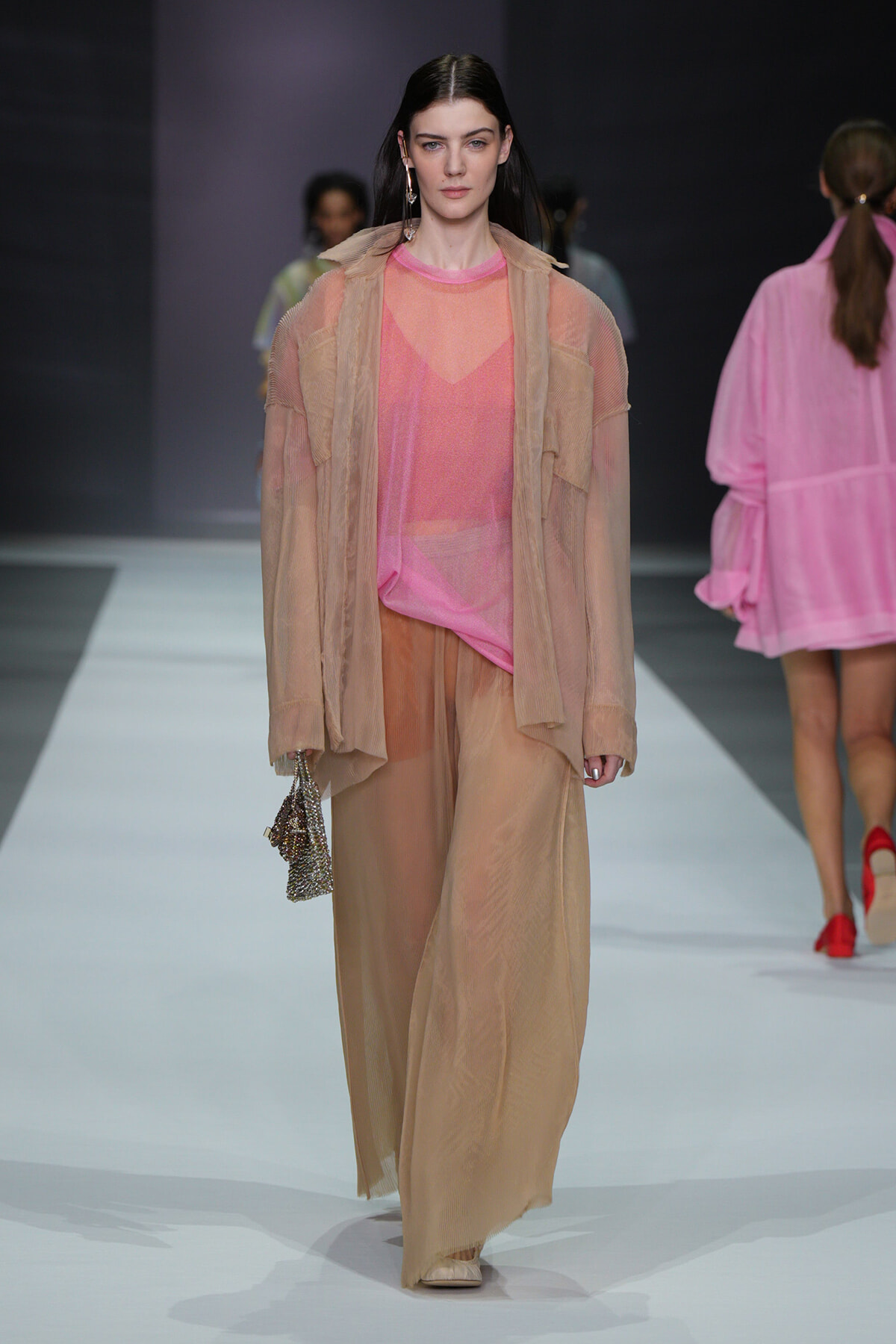 Model walks runway in peach sheer shirt and oversized beige outerwear with wide beige trousers, holding a beaded clutch.