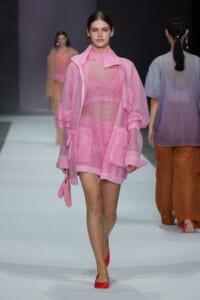 Model walking the runway in a pink mesh oversized jacket worn over a sheer pink top and matching mini dress, with a small pink bag and red flats.