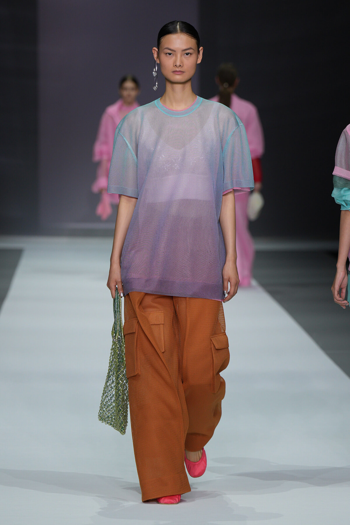 Model walking the runway in a pastel gradient oversized top (blue to purple) with orange cargo pants and pink shoes, carrying a green mesh bag.