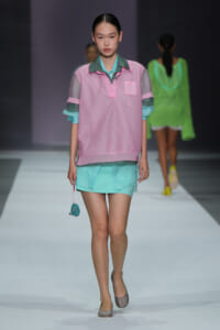 Model walking the runway in a pastel pink mesh polo over a turquoise shirt with mint-green shorts and a small teal beaded bag in hand.
