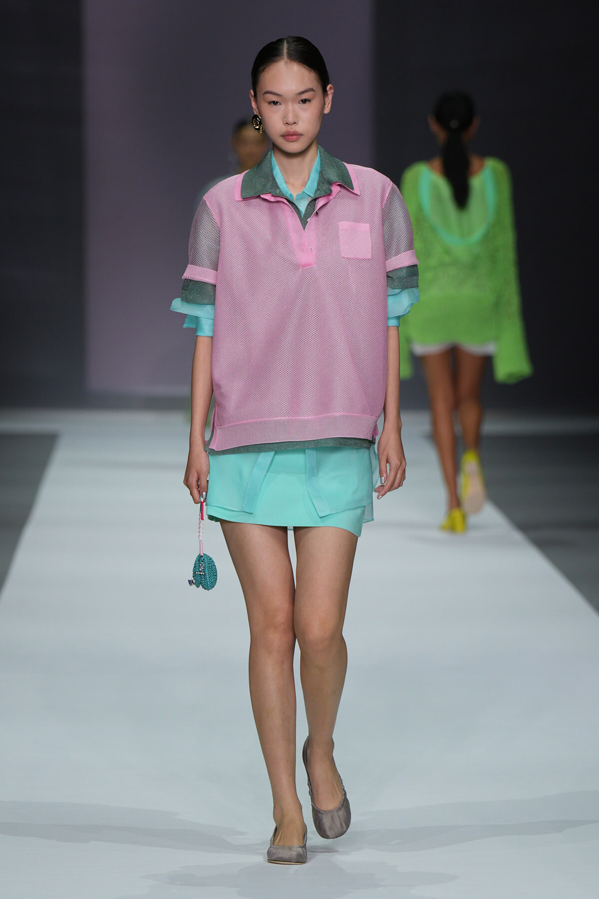 Model walking the runway in a pastel pink mesh polo over a turquoise shirt with mint-green shorts and a small teal beaded bag in hand.
